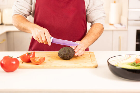 A man is cutting a tomato and an avocado on a cutting board. The man is wearing a red