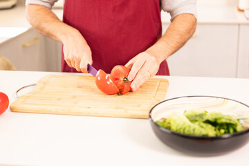 A man is cutting a tomato on a cutting board. The tomato is sliced in half and the knife is being used to cut it. The man is wearing a red apron