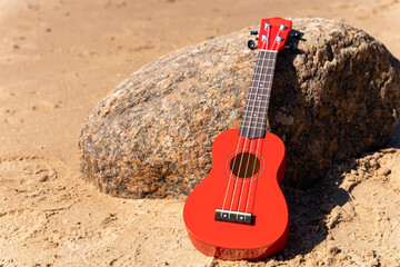 A red ukulele rests on a sandy beach under the sun