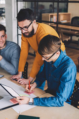 Group of male partners cooperating together in workspace sitting at desktop and making research via technology, young caucasian man reading information from internet and writing notes on stick