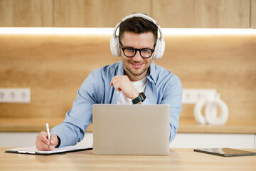 A focused young man wearing headphones works on a laptop while jotting notes, set in a contemporary...