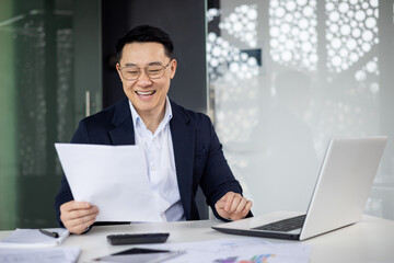 Joyful Asian businessman in glasses reviews successful reports at his modern office, displaying...