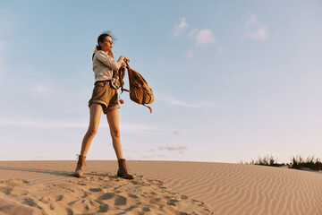 Female traveler standing triumphantly on sand dune with backpack, enjoying breathtaking desert...