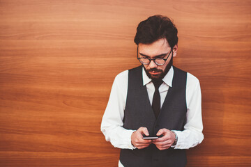 Young caucasian hipster guy in fashionable waistcoat chatting with followers in social networks using modern smartphone device and wireless internet near copy space area for advertising text
