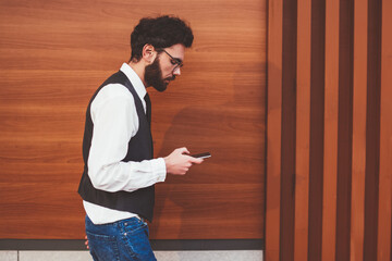 Trendy male blogger walking on publicity area and installing media application on cellular phone, young stylish hipster guy dialing number on modern smartphone device during free time outdoors