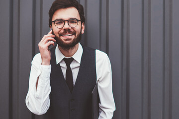 Half length portrait of cheerful hipster guy looking at camera during positive international telephone conversation outdoors, positive man in spectacles using roaming connection on publicity area