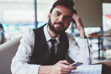 Portrait of positive hipster guy holding cellular phone in hands while resting at cafeteria during planning organisation, caucasian bearded male blogger enjoying time indoors using modern telephone