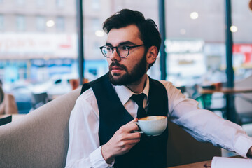 Contemplative caucasian hipster guy pondering on best seller holding cup with hot cappuccino at cozy cafeteria, young thoughtful man in spectacles looking away while sitting with textbook at table