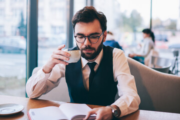 Concentrated hipster guy in eyeglasses for vision correction holding cup with hot tea during free time on leisure, handsome young male student enjoying caffeine beverage at university cafeteria