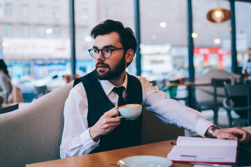 Contemplative caucasian hipster guy pondering on best seller holding cup with hot cappuccino at cozy cafeteria, young thoughtful man in spectacles looking away while sitting with textbook at table