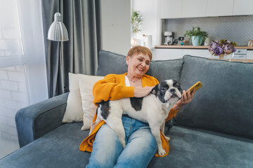 Elderly Caucasian woman hugging her pet French bulldog while sitting on the sofa at home, love for pets