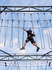 A teenage boy in sports safety equipment crosses a suspension rope bridge in a rope amusement park. View from below on the background of the sky