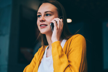 Positive charming hipster girl talking during telephone conversation on cellular sitting in cafeteria, beautiful woman calling on smartphone using good high speed connection enjoying coffee break