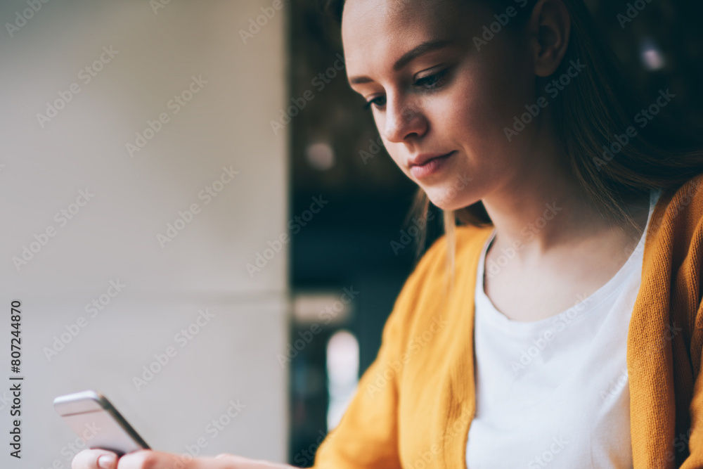 Poster concentrated female student sending sms message on smartphone while sitting at table with laptop com