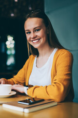 Happy caucasian hipster girl posing at cafe interior with cup of cappuccino during free time on leisure, cheerful woman in fashionable apparel looking at camera and enjoying coffee break indoors