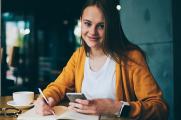 Portrait of successful female student doing homework and making notes in notepad reading information from internet websites using smartphone, cheerful hipster girls holding cellular and writing essay