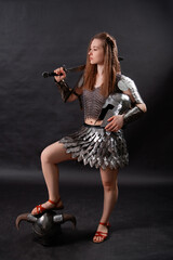 Full-length portrait of a medieval woman warrior in female armor with a sword in her hand standing in a heroic pose with her foot on a horned helmet isolated on a dark background.