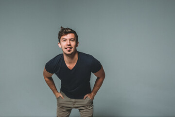 A man on a gray background in a dark blue T-shirt, hands in pockets, leans forward and shows his...