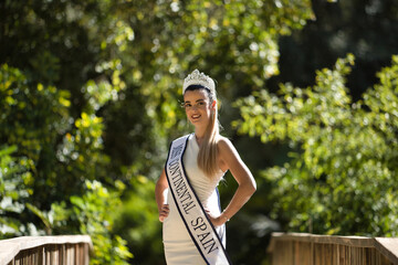 Young, beautiful, blonde woman in white suit, diamond crown and beauty pageant winner's sash, posing in the middle of a wooden bridge. Concept beauty, contests, pageant, fashion. - obrazy, fototapety, plakaty