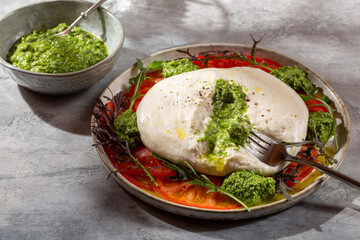 Tomato salad with burrata cheese and green pesto on gray background.