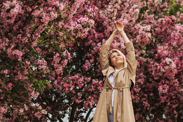 Relaxed blonde woman with arms raised up surrounded by blossoming apple trees in spring