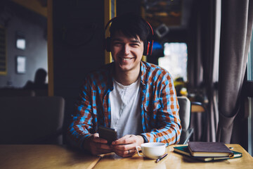 Happy male meloman listening favourite music in stylish modern headphones while holding smartphone in hands sitting at wooden table with cup of tasty coffee and books in coworking space