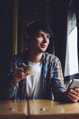 Handsome hipster guy with cup of coffee in hand enjoying music via headphones.Smiling young man with modern mobile phone listening playlist in earphones while looking out of window