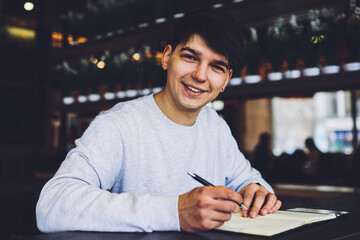 Half length portrait of handsome skilled student with pen in hand smiling at camera while preparing...