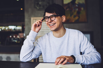 Positive young man in spectacles with good lenses remembered funny case of life for writing interesting story sitting at coffee table in stylish cafeteria.Cheerful student laughing during homework