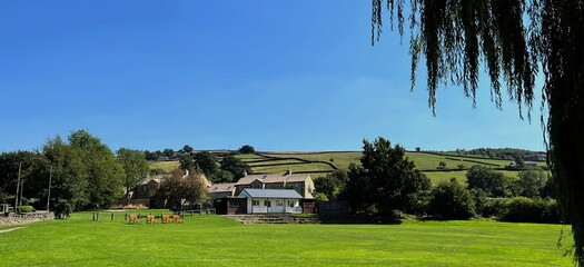 A serene landscape, with a lush green field and a small cricket pavilion, in the background are  rolling hills and a clear blue sky in, Bradley, Keighley, UK