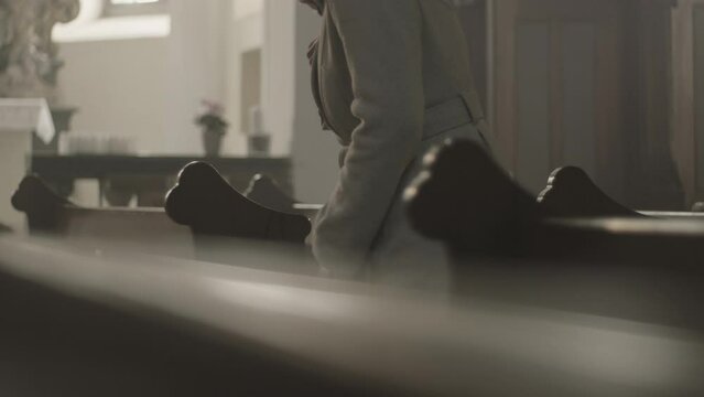 Religious woman crossing herself and sitting on pew bench in Christian church side view medium shot. Mid adult caucasian person kneeling and praying in cathedral