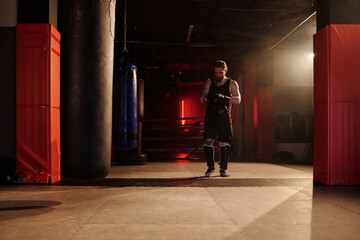 Long shot of young male athlete in black sportswear walking along gym with punching bags and...