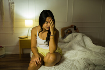 Latin woman holding her head in discomfort while a man lies in the background, depicting tension or headache