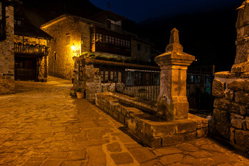 An old stone fountain on a quiet cobblestone street. The street is lit up with a yellow light at...