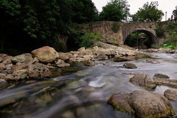 Long exposure view of a river with an old stone bridge over it and rocks in the water and shores, . Barcena Mayor, Saja-Besaya Natural Park, Cantabria, Spain