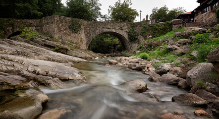 Panoramic view of an old stone bridge over a river. Long exposure view of the calm waters on an overcast day, Barcena Mayor, Saja-Besaya Natural Park, Cantabria, Spain.