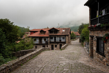An old small village with cobblestone streets and traditional houses with red tiles roofs with foggy mountains in the background. Barcena Mayor, Saja-Besaya Natural Park, Cantabria, Spain