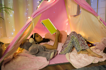 Little girl finds comfort and joy reading a book in a homemade tent with fairy lights in her bedroom at night