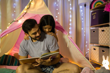 Father and daughter enjoy a bedtime story in a homemade tent adorned with fairy lights, creating a warm nightly ritual