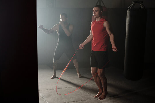 Light falling from window of gym on two young male athletes practicing jumps with skipping rope and boxing techniques with punching bag