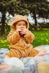 Cute baby girl in plush suit with red apple in hands sitting on blanket in green grass.