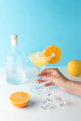 Tropical cocktail in martini glass with ice cubes and bottle of alcohol on white table. Minimal summer drink composition.