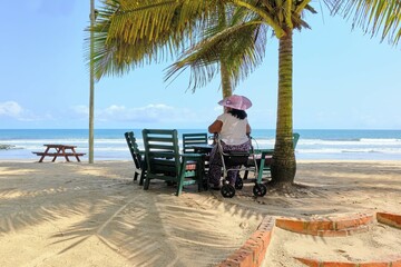 Senior African American woman sitting on a mobile rollator walker under palm trees at golden sandy...