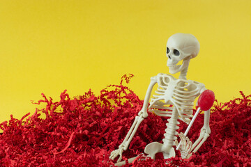 Skeleton holding cherry lollipop sitting in red shredded crinkled paper against yellow sky