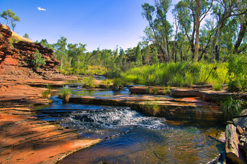 Red rocks, a clear stream with a cascade, and lush green vegetation in the Kalamina Gorge, Karijini...