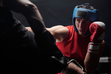Boxer with protective shield on head keeping left hand in boxing glove by face and looking at rival...