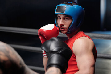 Young muscular man in red vest and boxing gloves and blue face shield looking at his rival before...