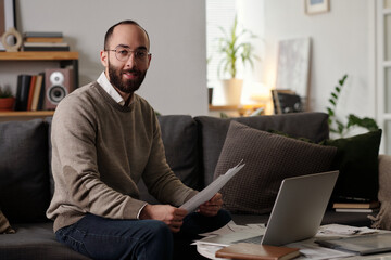 Smiling bearded man with financial documents in hands looking at camera while sitting on couch in...