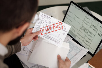 High angle of young man holding envelope and taking out financial bill stamped as past due while sitting by table with other documents