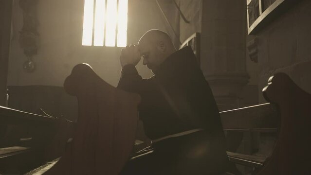 Monk in robe praying on pew in church side view medium shot. Christian clergyman with closed eyes and clasped hands sitting on wooden bench in monastery